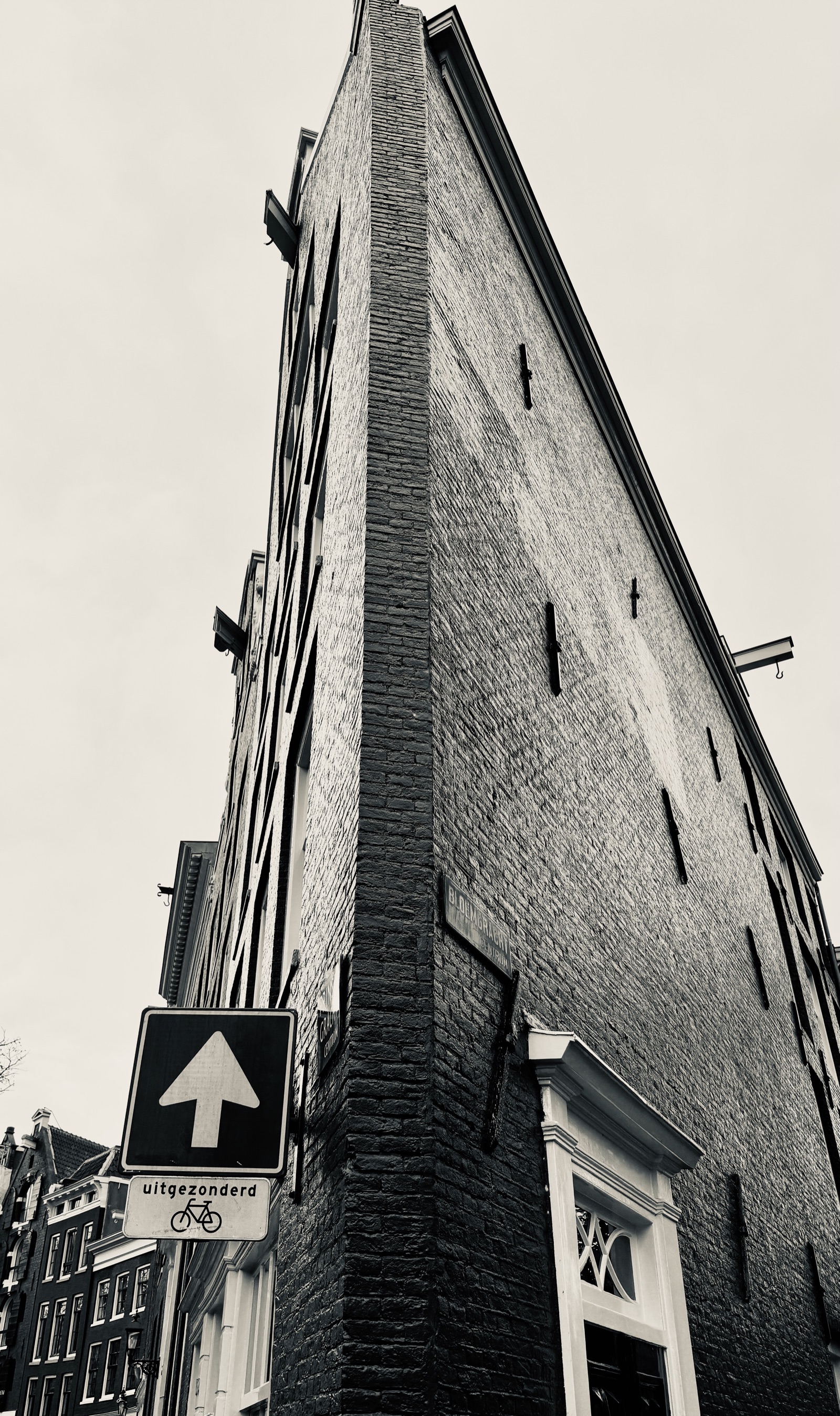 Giebel — Ein schmales Amsterdamer Giebelhaus, steil von unten fotografiert, dunkler Backstein gegen den bleichen Himmel · Randlicht Fotografie